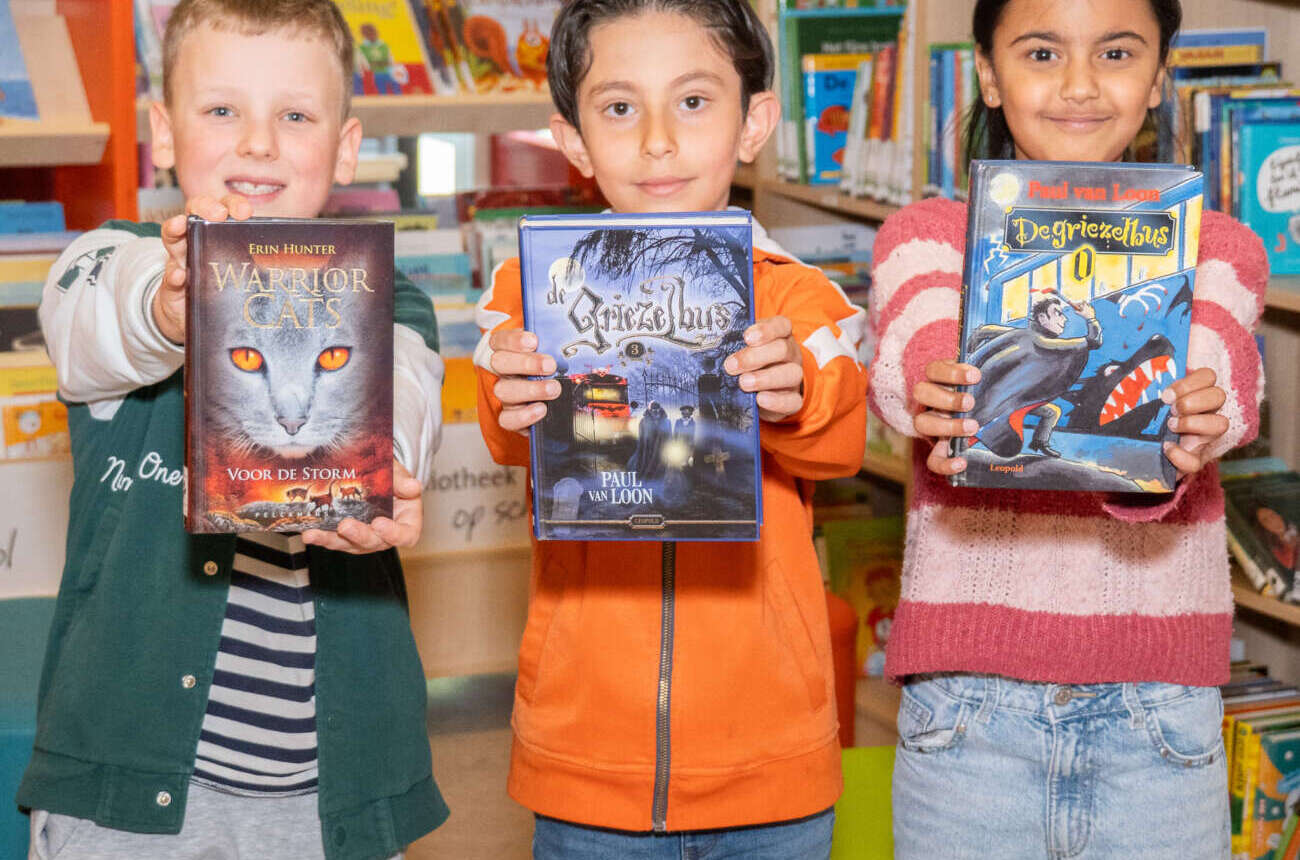 Drie leerlingen tonen trots hun favoriete boeken in de schoolbibliotheek van De Regenboog