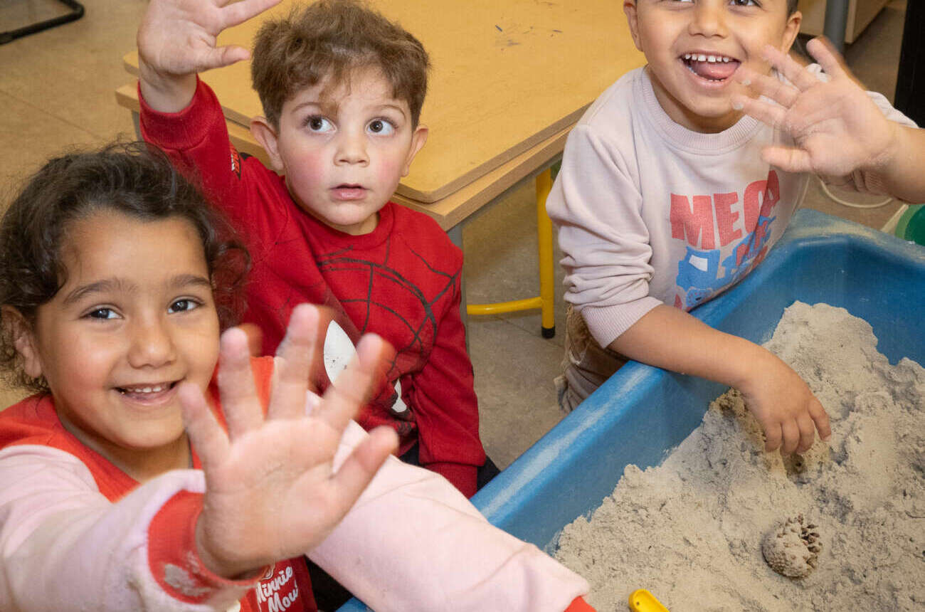 Drie lachende kleuters spelen samen in de zandtafel en zwaaien naar de camera