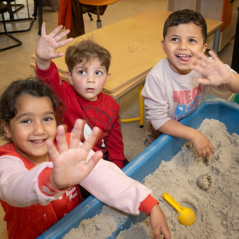 Drie lachende kleuters spelen samen in de zandtafel en zwaaien naar de camera