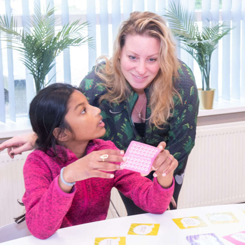 Leerkracht buigt zich vriendelijk over een leerling die werkt met taalkaartjes
