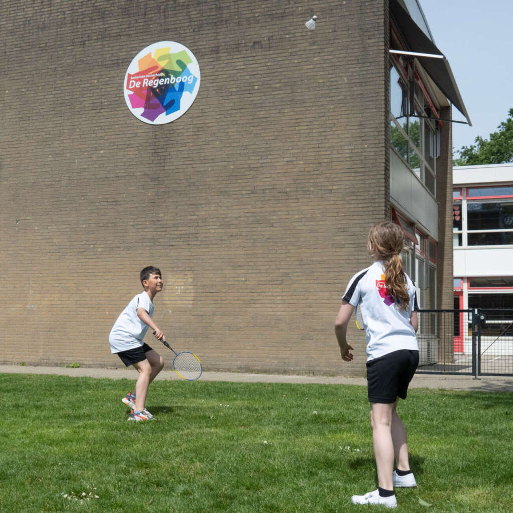 Twee leerlingen in shirts van De Regenboog spelen badminton op het gras voor het schoolgebouw