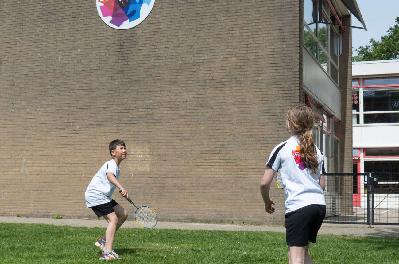 Twee leerlingen in shirts van De Regenboog spelen badminton op het gras voor het schoolgebouw