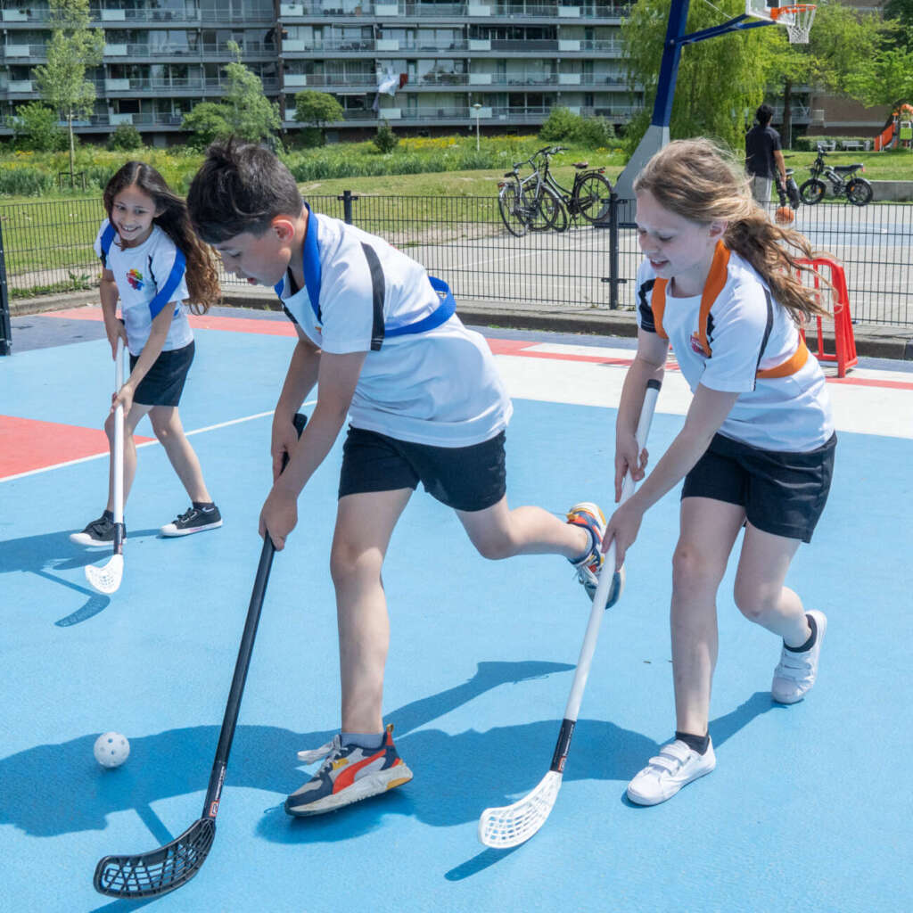 Drie leerlingen spelen unihockey op het blauw-rode sportveld bij de school
