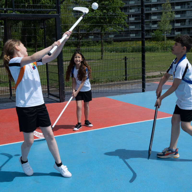 Drie enthousiaste leerlingen spelen unihockey in de buitensportkooi van De Regenboog