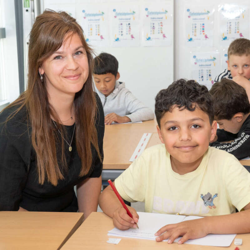 Lachende leerkracht zit naast een leerling die geconcentreerd schrijft in een schrift