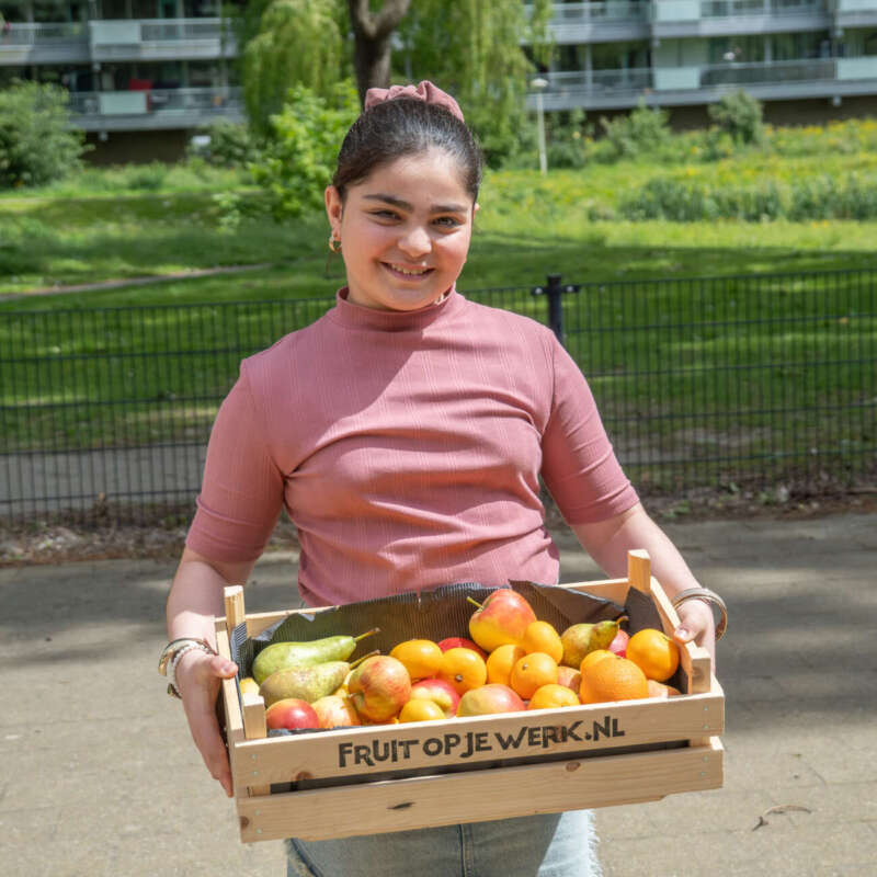 Glimlachende medewerker draagt een krat vers fruit van Fruit op je werk de school in