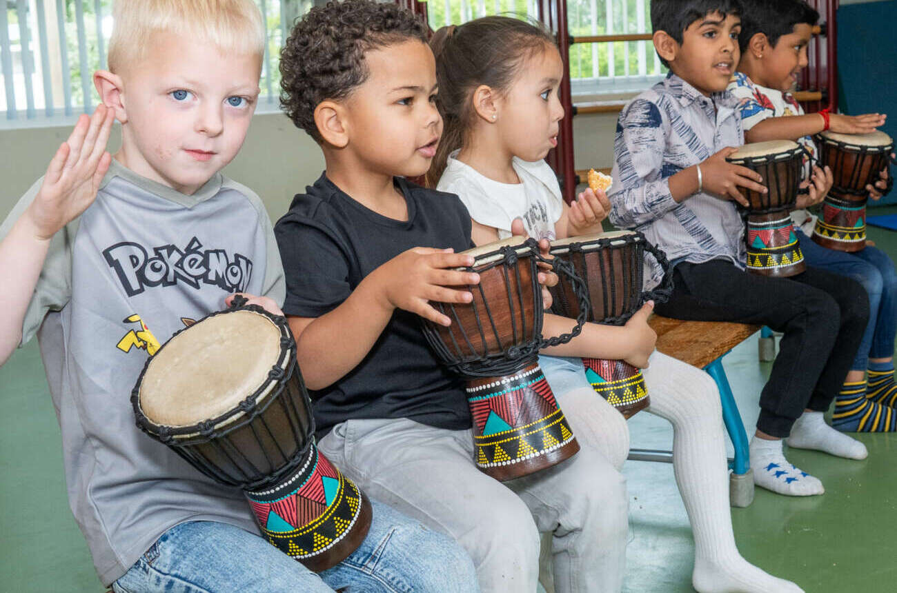 Vijf kleuters spelen samen op djembés tijdens een muziekles in de gymzaal