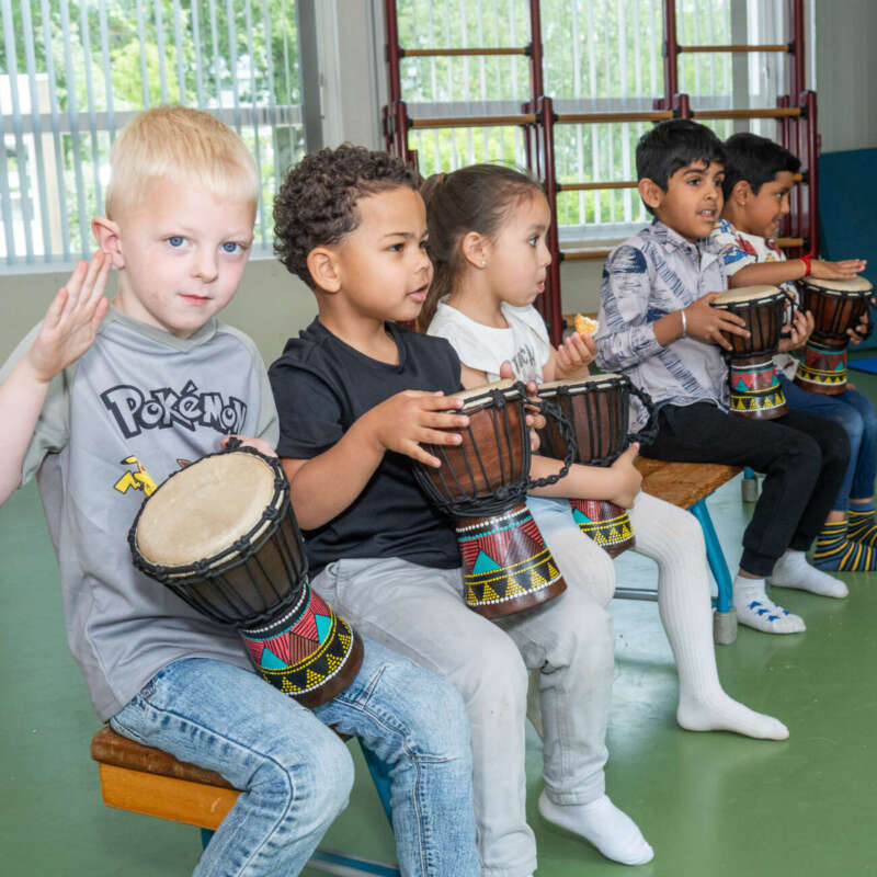 Vijf kleuters spelen samen op djembés tijdens een muziekles in de gymzaal